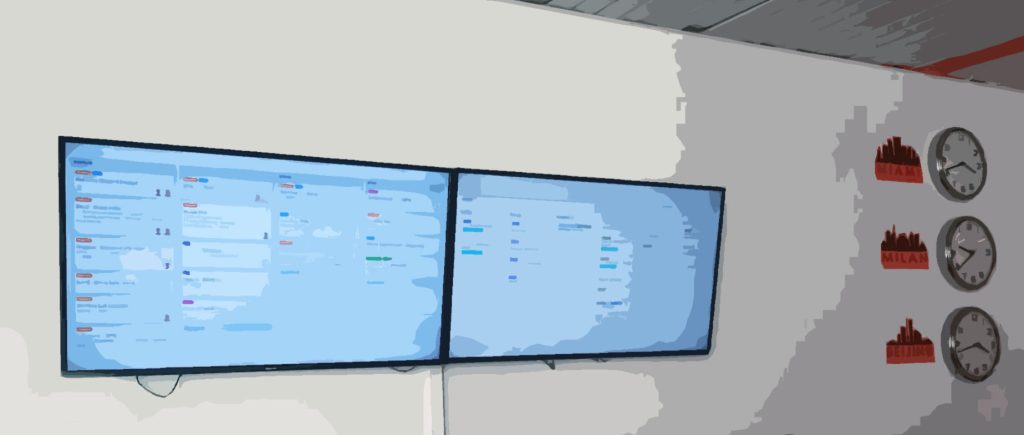 due schermi con board organizzative appesi su parete sopra ad una postazione di lavoro